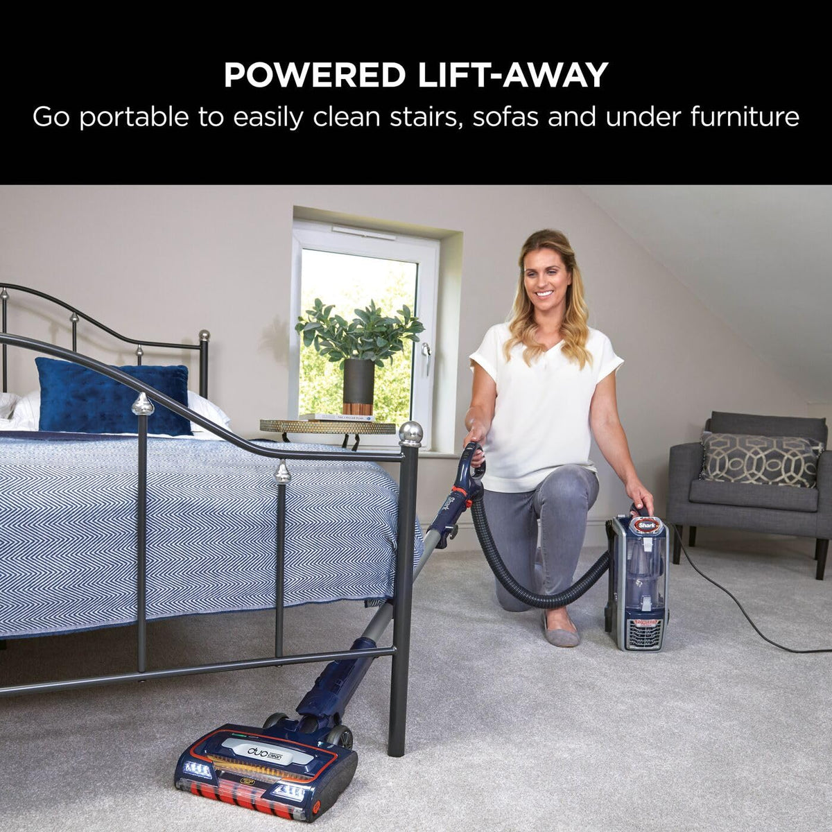 Woman using the Lift-Away portable canister and hose to vacuum a bedroom floor beside a bed.
